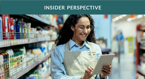 A woman grocery store employee in the kitchen supply aisle looking over data on a tablet.
